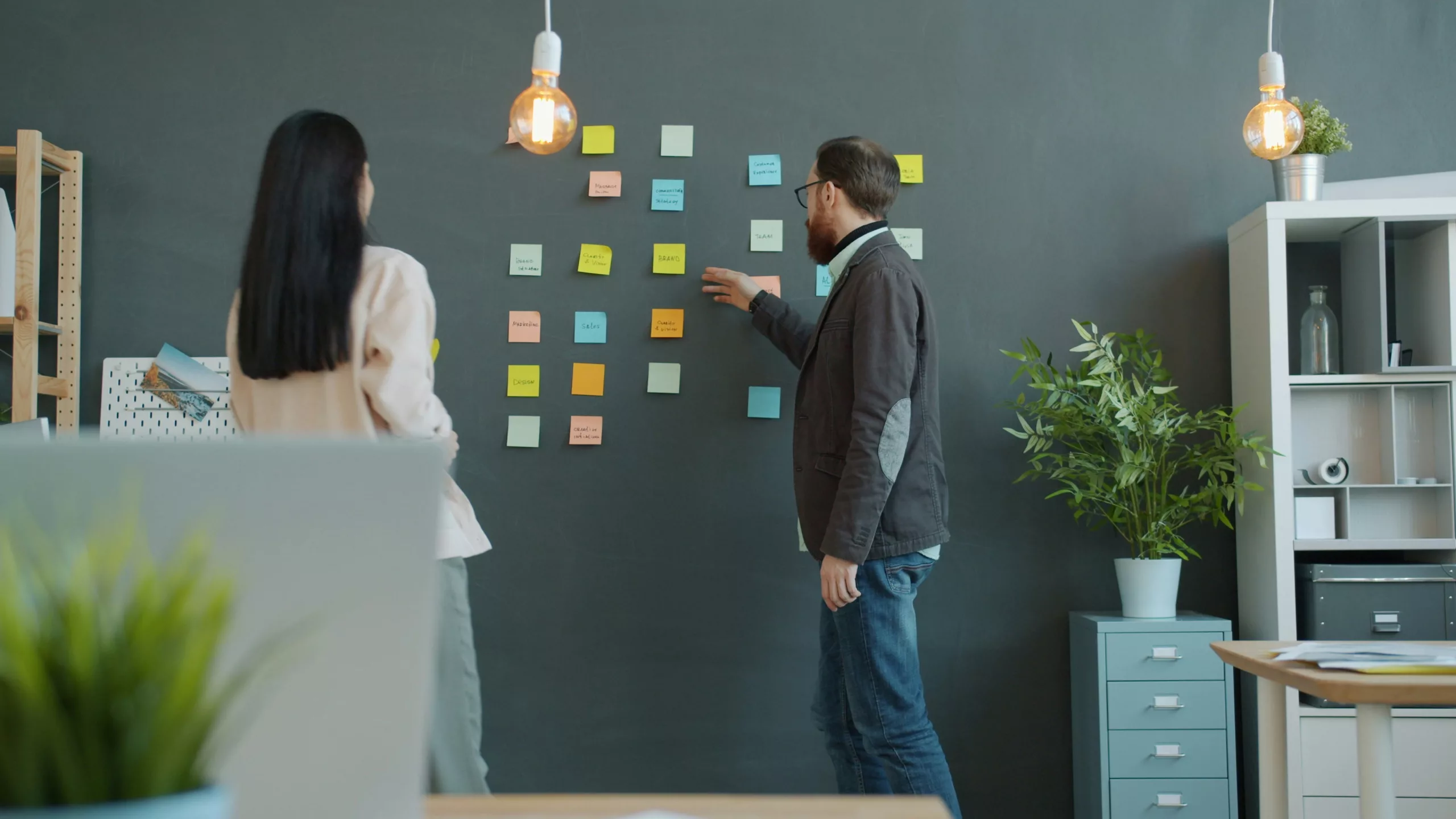 Dos personas haciendo el benchmarking, analizan información de la competencia utilizando notas adhesivas organizadas por categorías en una pared de pizarra, en un entorno de oficina moderno.